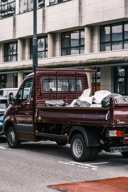 A small maroon flatbed truck is parked on a city street in front of a modern office building with large glass windows and a concrete facade. The truck's cargo area is filled with various discarded items wrapped in grey and white plastic bags, some appearing bulged and loosely tied, indicating they contain waste or unwanted materials. A metal safety grille is attached to the cab's rear side, providing added security. The pavement shows clear lane markings, and nearby vehicles are partially visible, suggesting an urban environment suitable for private rubbish removal or on-site clearance services. The scene is illuminated by natural daylight, with the building reflecting the surrounding urban landscape. The presence of the truck and waste materials aligns with independent collection or alternative waste handling options often offered by companies like Rubbish Clearance Haringey, emphasizing their role in handling bulk waste removal from commercial or residential premises without reliance on local authority services.