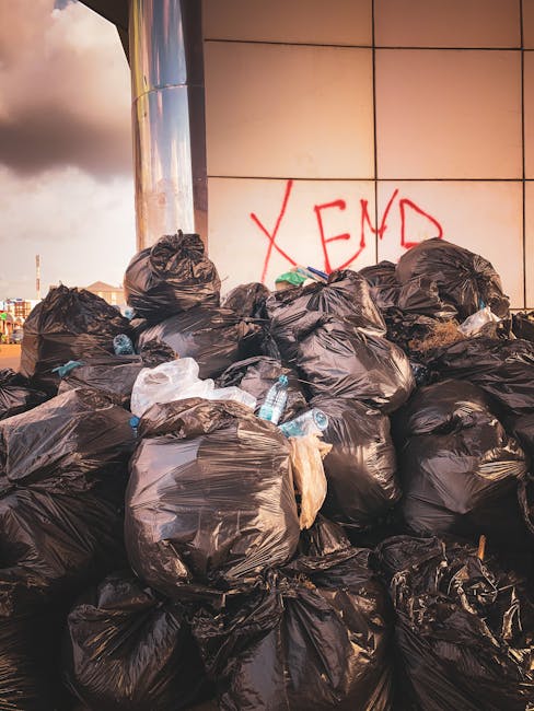 A large pile of black plastic rubbish bags filled with waste is stacked against a modern building with a tiled exterior. Some bags are partially torn, revealing plastic bottles and other debris inside. The bags vary in size and are tightly knotted at the top, with a few white and clear plastic bottles visibly protruding or lying on the ground among the bags. Behind the pile, there is graffiti spray-painted in red on the wall, spelling 'XEND.' The scene appears to be outdoors on a paved surface, likely in an urban setting, with cloudy sky visible in the upper left corner. The overall environment suggests an area awaiting removal by a waste disposal service like Rubbish Clearance Haringey, reflecting a typical scenario of excess rubbish accumulated for private rubbish collection or alternative waste handling outside local authority services.
