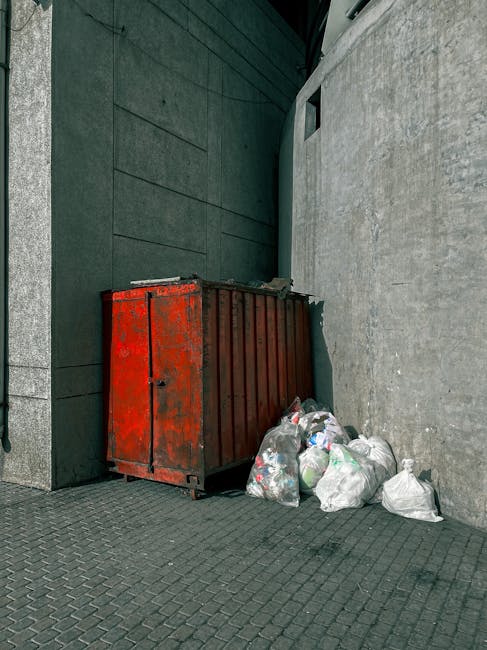A pile of black rubbish bags, cardboard boxes, and discarded packaging materials are stacked against a weathered, beige exterior wall near a small, red metal door that is covered in graffiti and secured with a brass handle. The debris appears to be gathered from an indoor or outdoor space, likely awaiting removal, with some items partially smashed or torn. Visible to the left side are additional excluded rubbish items, including plastic containers and boxes, indicating a collection of household or commercial waste in need of proper disposal. The wall's rough texture and the surrounding urban environment suggest an informal rubbish accumulation site, typically associated with independent waste clearance services such as those offered by Rubbish Clearance Haringey, which provide alternative waste handling solutions outside traditional municipal collections. The scene reflects an area where rubbish is temporarily stored before professional removal, emphasizing the importance of regulated rubbish clearance for maintaining cleanliness and compliance with local waste disposal guidelines.
