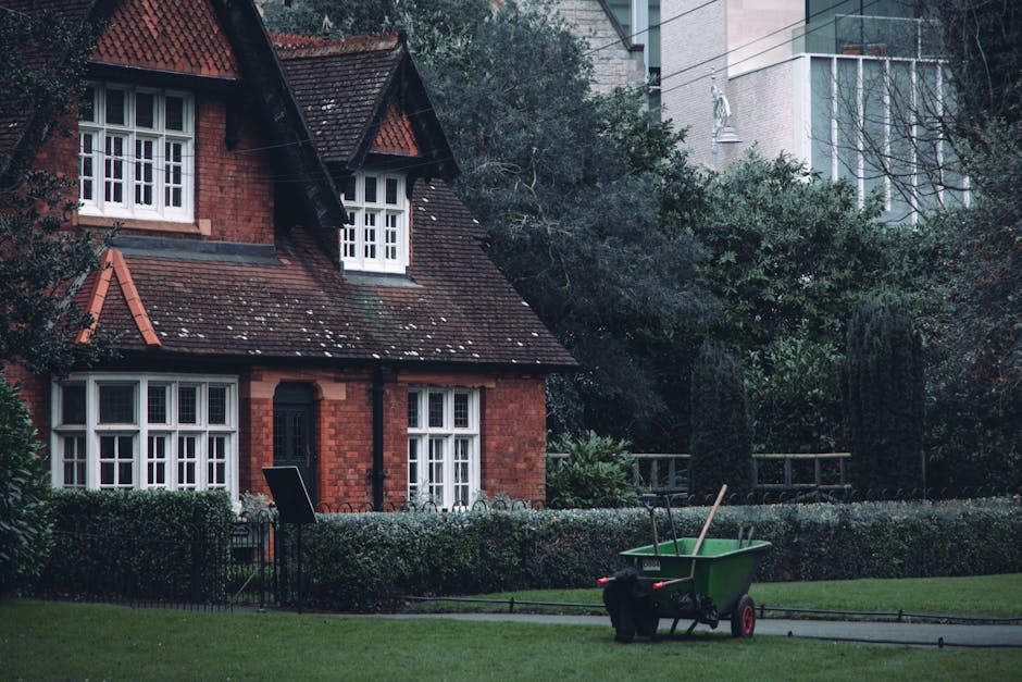 A green wheelbarrow with red wheels and black handles is positioned on a paved pathway in front of a well-maintained garden. The wheelbarrow contains a garden fork with a wooden handle and a metal rake, alongside a black bag. Surrounding the pathway is a neatly trimmed hedge with dense, dark green foliage. Behind the hedge, there is a red-brick house with a dark tiled, steeply pitched roof featuring multiple white-framed bay windows and a small overhanging dormer window. The house is set within a residential area with a lush garden environment, including several mature trees and shrubs. In the background, parts of modern high-rise buildings with glass facades are visible, indicating an urban setting close to the countryside. The weather appears overcast, with diffuse lighting illuminating the scene evenly, suggesting a typical day suited to ongoing outdoor garden maintenance, which occasional private waste collection or rubbish clearance services like Rubbish Clearance Haringey might support through on-site clearance or alternative waste handling solutions.