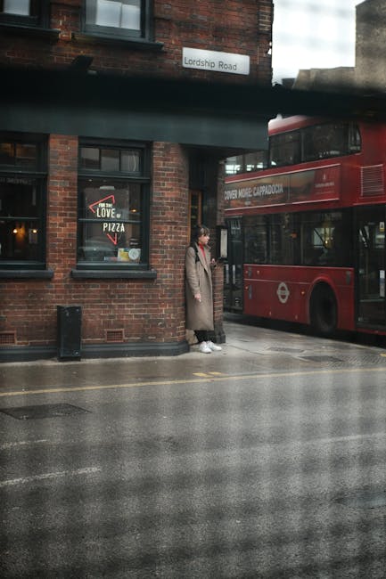 A woman standing on a damp, reflective pavements outside a red double-decker bus parked along a brick building with dark window frames, situated on Lordship Road. The woman wears a long beige coat, white sneakers, and carries a black handbag. The building features a window with a pizza advertisement and a sign displaying the street name. The scene appears to be in an urban area with a cloudy sky, and the bus is part of the London Transport fleet. The environment suggests a typical city street, possibly during or after rain, with a mix of commercial and residential surroundings. This setting aligns with locations where independent waste collection or private rubbish removal services like those offered by Rubbish Clearance Haringey might operate to manage local waste and bulky items, providing an alternative to council-based rubbish removal methods.