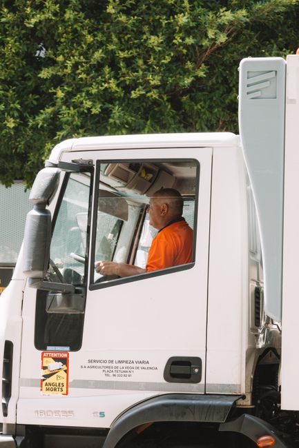 Close-up side view of a white waste collection truck with the driver visible through the window, an individual wearing an orange high-visibility vest seated inside. The truck's exterior features safety and warning stickers, including an orange warning sign with the word 'ATTENTION' and hazard symbols. The background includes dense green foliage and trees, indicating an outdoor setting likely near a residential or commercial property. The vehicle is parked on a paved surface, and the scene suggests it is part of an independent rubbish clearance service, typical of private waste handling operations, in alignment with rubbish removal activities. The image captures the professional and functional nature of the vehicle used by Rubbish Clearance Haringey for doorstep or on-site refuse collection, emphasizing safety and operational readiness in waste management.