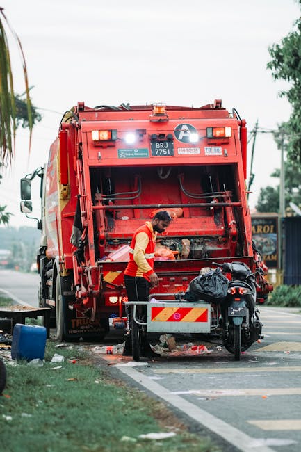 A street scene showing a worker from Rubbish Clearance Haringey using a red waste collection vehicle for rubbish removal. The worker is dressed in an orange high-visibility vest over a yellow long-sleeved shirt and is positioned at the rear of the truck, handling black rubbish bags placed on a small metal platform. The waste collection vehicle prominently features a large, red, industrial metal body with a textured, painted surface and reflective safety markings on its rear. The vehicle's compartment is open, revealing mechanical components and cleaning tools inside. The scene takes place on a paved urban street with a curb and a defined roadside area, including some discarded rubbish on the ground nearby and a blue container placed on the grass. In the background, there are trees, utility poles, and buildings, and the environment appears overcast with natural daylight illuminating the area. The scene exemplifies a professional, independent waste disposal activity aligned with private rubbish clearance services, emphasizing on-site collection and rubbish handling outside of municipal services.