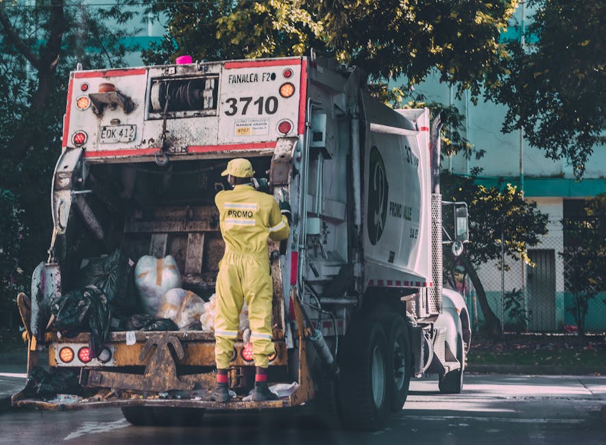 Close-up side view of a white waste collection truck with the driver visible through the window, an individual wearing an orange high-visibility vest seated inside. The truck's exterior features safety and warning stickers, including an orange warning sign with the word 'ATTENTION' and hazard symbols. The background includes dense green foliage and trees, indicating an outdoor setting likely near a residential or commercial property. The vehicle is parked on a paved surface, and the scene suggests it is part of an independent rubbish clearance service, typical of private waste handling operations, in alignment with rubbish removal activities. The image captures the professional and functional nature of the vehicle used by Rubbish Clearance Haringey for doorstep or on-site refuse collection, emphasizing safety and operational readiness in waste management.