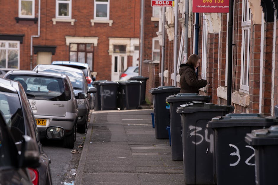 A woman standing on a residential pavement beside several black plastic wheelie bins, which are lined up along the front of a row of red-brick terraced houses. The bins have white painted numbering and are being used for household waste disposal. The street features parked cars, including a visible silver hatchback closest to the foreground, with additional vehicles further down the street. The houses have sash windows with white frames, some with small window sills, and the facades show signs of typical brickwork with contrasting white stone lintels. Overhead, a red sign for a property sale is partially visible, attached to a lamppost, indicating the presence of real estate activity. The scene appears to be in natural daylight with an overcast sky, providing even lighting across the street. This setting reflects typical urban waste management, where privately arranged rubbish collection services like those offered by Rubbish Clearance Haringey may be used for alternative waste removal and on-site clearance, especially before property changes or cleanouts.