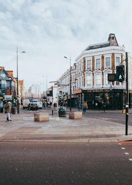 A busy street scene featuring a corner building with classic white and brown brickwork, housing The World's End pub, with large windows and decorative architectural details. The street is paved with large, rectangular stone tiles, and a pedestrian crossing with traffic lights and a green walking signal is visible in the foreground. Several pedestrians are walking across and along the pavement, some wearing coats. To the left, there are parked cars and a delivery van driving in the background, with more vehicles further along the street. On the right, there are small planters with no visible plants, positioned on the edge of the pavement near the crossing. The sky is overcast, contributing to a diffused, natural light. This urban scene relates to the context of independent collection and private waste handling, characteristic of an environment where rubbish clearance services, such as those offered by Rubbish Clearance Haringey, might operate in areas with high pedestrian activity and street-level commerce.