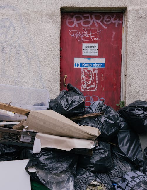 A pile of black rubbish bags and assorted discarded items, including cardboard and plastic, are gathered on the pavement in front of a red metal door covered with graffiti and stickers, located on an external wall with a rough, textured surface. The bags appear to contain household or general waste and are stacked haphazardly, some leaning against the wall. The door has signs attached, one indicating 'No Money' and another with instructions to 'Keep Clear,' suggesting this is an area dealing with private waste disposal rather than official refuse collection. The surrounding environment appears to be an urban setting, possibly an alleyway or back street, with limited natural light illuminating the scene. This image reflects the work involved in independent rubbish clearance services, such as those provided by Rubbish Clearance Haringey, which handle excess waste left outside properties, supporting alternative disposal methods outside of council collection systems.