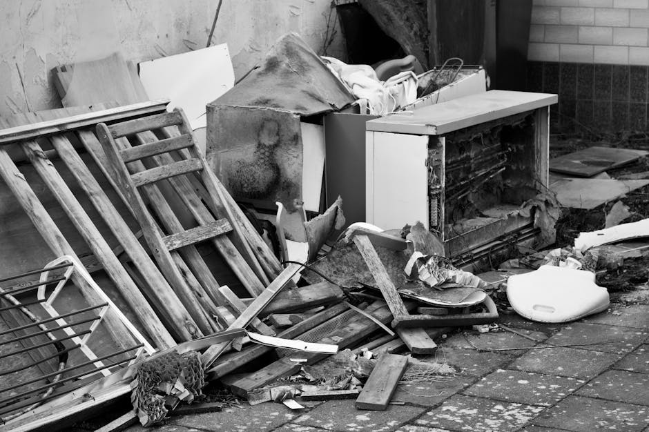A collection of discarded household waste and debris piled on a paved outdoor area next to a brick wall. The image features a weathered wooden bed frame with slatted sides leaning against a wall, alongside a partially disassembled cardboard box filled with miscellaneous objects. Visible materials include wood, metal, plastic, and fabric, with textures ranging from rough and splintered to smooth and shiny. There is a large, irregularly shaped piece of discarded furniture or insulation that appears stained and worn, resting atop the pile. Scattered around are fragments of broken wood, plastic sheets, and torn fabric, along with a white foam or plastic object resembling a seat or cushion. The scene is in natural daylight, highlighting the contrast between the various materials and weathered surfaces. This image illustrates the type of bulk waste collection that companies like Rubbish Clearance Haringey handle through private disposal or on-site clearance, providing an alternative to municipal rubbish collection and demonstrating the diverse waste types often involved in junk removal services for properties in the area.
