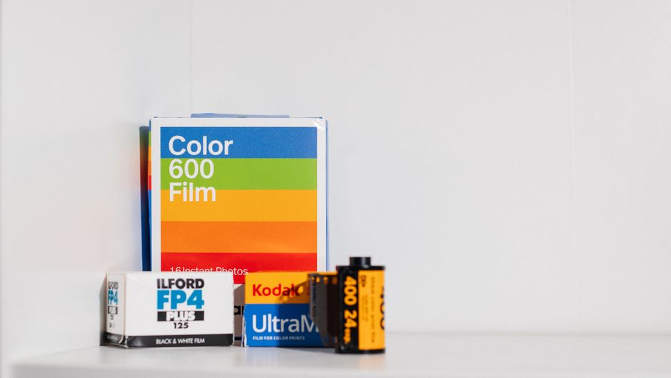 A close-up view of a collection of photographic and film-related items arranged on a white surface against a plain, light gray background. The backdrop features various boxes, including a larger rectangular one labeled 'Color 600 Film' in white text with a colorful striped design, positioned toward the left side of the image. In front of this, there is a smaller rectangular box labeled 'Ilford FP4 Plus 125' in black, gray, and blue, situated slightly toward the left. To the right, overlapping the film boxes, is a vintage Kodak Ultra Max 400 24 exposure 35mm film roll with a distinctive orange and black design, and beside it, an orange and black 400 24 exposure film roll with a plastic core. The items showcase a mix of textures: glossy and matte finishes on the boxes, and the smooth plastic of the film canisters. The overall composition illustrates equipment related to traditional film photography, emphasizing the materials, colors, and packaging typical of on-site or private photography material storage, reflecting a context of alternative media handling or collection for reuse or disposal by companies such as Rubbish Clearance Haringey.