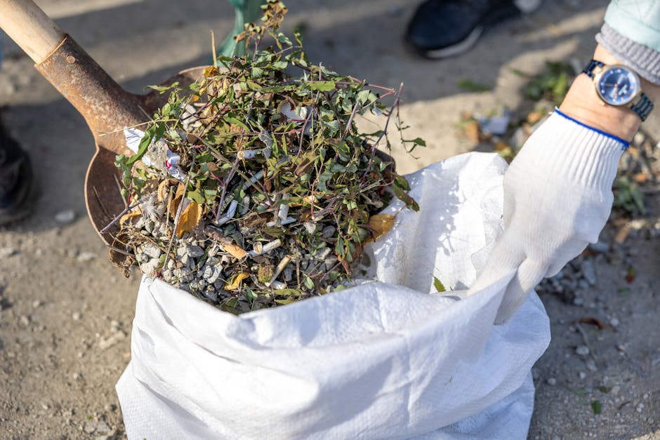 A person wearing a white glove with blue trim and a silver wristwatch is holding open a white, woven polypropylene bag filled with garden debris, primarily consisting of small branches, green leafy plant material, and a mix of soil and gravel. The debris includes twigs with variegated green and purple leaves, some dried brown leaves, and small stones, all piled loosely in the bag. In the background, part of a rusted metal shovel handle is visible, positioned at an angle above the bag, indicating the material may have been recently collected or is in the process of being gathered for removal. The scene takes place outdoors on a dirt or gravel surface, with a person's dark shoe visible on the right side, suggesting a site where waste or garden clearance is being carried out by a waste management service such as Rubbish Clearance Haringey, who specialise in private waste disposal and alternative collection methods outside local authority services. The lighting appears natural, casting soft shadows on the ground, providing a clear view of the mixed natural and man-made materials being prepared for disposal in an environment typical of yard or garden clearance work.
