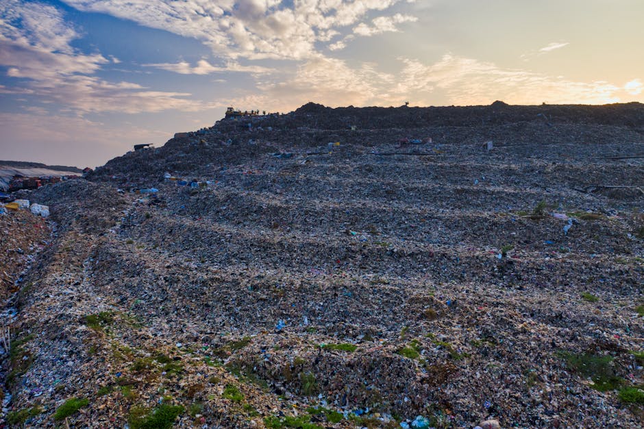 A large landfill site during late afternoon or early evening, showing a vast area layered with dark, crushed rock and compacted waste materials. The terrain is uneven with visible mounds and slopes, with small scattered patches of green vegetation growing sporadically across the site. In the background, the landfill extends up toward the horizon, where a group of silhouetted figures or structures can be seen at the top, beneath a sky filled with a mix of scattered clouds and warm, fading sunlight. The scene demonstrates the excess waste accumulation and site management typical of private waste disposal operations and highlights the importance of professional rubbish removal services, such as those provided by Rubbish Clearance Haringey, which may include the clearance of such large waste deposits or construction debris from sites needing on-site or independent collection solutions.