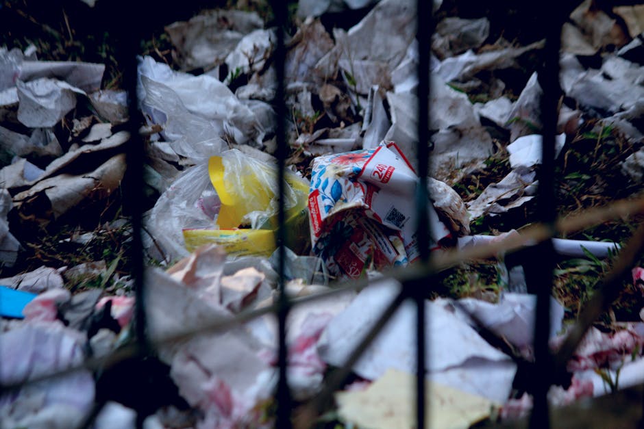 A person wearing a white glove with blue trim and a silver wristwatch is holding open a white, woven polypropylene bag filled with garden debris, primarily consisting of small branches, green leafy plant material, and a mix of soil and gravel. The debris includes twigs with variegated green and purple leaves, some dried brown leaves, and small stones, all piled loosely in the bag. In the background, part of a rusted metal shovel handle is visible, positioned at an angle above the bag, indicating the material may have been recently collected or is in the process of being gathered for removal. The scene takes place outdoors on a dirt or gravel surface, with a person's dark shoe visible on the right side, suggesting a site where waste or garden clearance is being carried out by a waste management service such as Rubbish Clearance Haringey, who specialise in private waste disposal and alternative collection methods outside local authority services. The lighting appears natural, casting soft shadows on the ground, providing a clear view of the mixed natural and man-made materials being prepared for disposal in an environment typical of yard or garden clearance work.